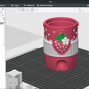 May include: A 3D digital model of a cylindrical container on a build plate. The container is pink with white polka dots, featuring a central white band and a strawberry emblem with green leaves and a white flower. A rectangular color swatch is nearby.