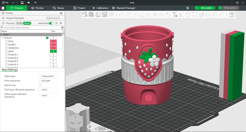 May include: A 3D digital model of a cylindrical container on a build plate. The container is pink with white polka dots, featuring a central white band and a strawberry emblem with green leaves and a white flower. A rectangular color swatch is nearby.
