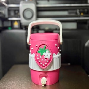 May include: A cylindrical pink beverage dispenser with a white handle and a white spigot at the base. The exterior features a raised strawberry emblem with a green leaf and a white flower, accented by small white dots and a white band around the middle.