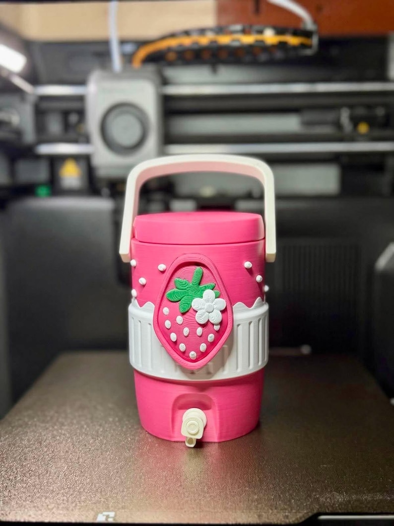 May include: A cylindrical pink beverage dispenser with a white handle and a white spigot at the base. The exterior features a raised strawberry emblem with a green leaf and a white flower, accented by small white dots and a white band around the middle.