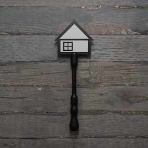 May include: A black and white house-shaped sign with a window, mounted on a black pole. The sign is set against a weathered, wooden background, creating a contrast of textures and colors. The design is simple and modern.