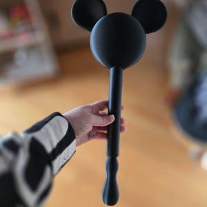 May include: A black decorative wand with a Mickey Mouse head design. The wand features a large circular head with two smaller circular ears on top. The handle is long and cylindrical, with a decorative bulbous end.
