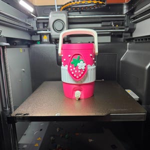 May include: A bright pink cylindrical beverage cooler with a white handle and a white spigot at the base. The exterior features a raised strawberry motif with a green leaf and a small white flower, accented by white dots and a white band around the middle.
