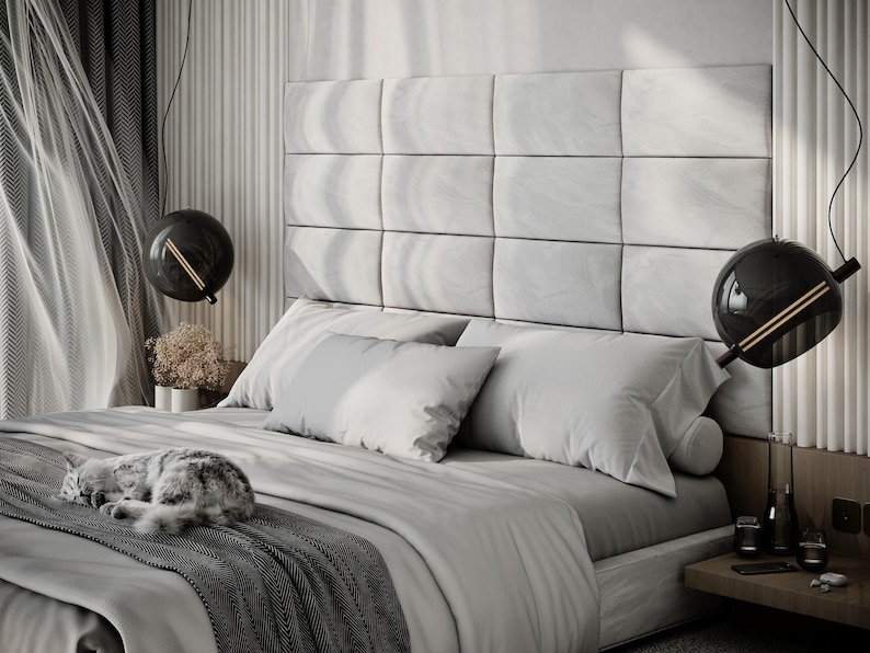 May include: A modern bedroom with a large bed featuring a gray headboard and bedding. Two spherical black pendant lights hang above the bed. A gray and white cat is curled up on the bed. The room has a neutral color palette.