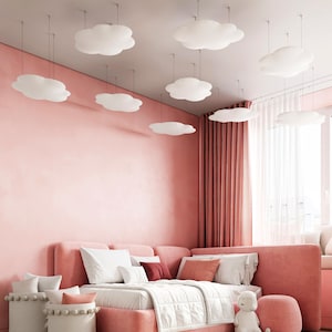 May include: A pink bedroom with cloud-shaped ceiling lights. The room features a large, plush pink sofa with white and red pillows, a white bedspread, and a matching ottoman. White baskets with pillows are in the foreground, and a window with curtains is in the background.
