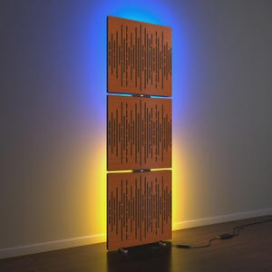 May include: A modern, vertical sound panel composed of three rectangular wooden sections. Each section features a pattern of vertical slits. The panel is illuminated with blue and yellow LED lighting, creating a gradient effect. A black cable extends from the base.