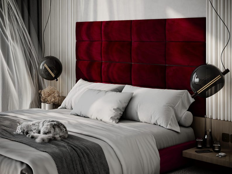 May include: A luxurious bedroom scene featuring a bed with a deep red, tufted headboard. The bed is made with white bedding and gray accents. A gray and white cat is curled up on the bed. Modern black globe pendant lights hang above.
