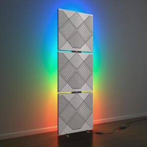 Könnte beinhalten: Eine moderne Leuchte mit drei weißen, perforierten Paneelen, die vertikal angeordnet sind. Die Paneele strahlen ein farbenfrohes Regenbogenlicht an die Wand. Ein schwarzes Netzkabel ist zu sehen.