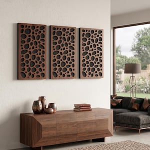 May include: Three rectangular wooden wall panels with a dark brown finish and a pattern of irregular, organic-shaped cutouts. The panels are mounted on a white wall above a wooden cabinet. Decorative vases and books are on the cabinet.