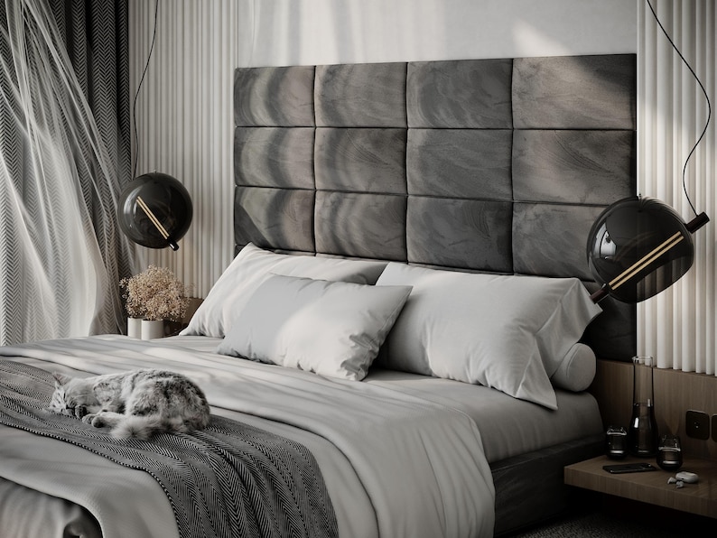 May include: A modern bedroom with a large bed featuring a gray headboard composed of square panels. The bed is covered with white and gray bedding, and a gray cat is curled up on the bed. Two black globe pendant lights hang above the bed.