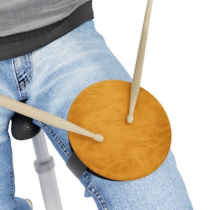 Könnte beinhalten: Ein hellbraunes Schlagzeug-Pad ist mit einem schwarzen Riemen am Knie einer Person befestigt. Zwei hölzerne Drumsticks sind über dem Pad positioniert, was auf die Verwendung hindeutet. Die Person trägt eine Jeans.