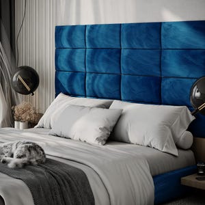 May include: A modern bedroom featuring a bed with a large, square, blue velvet headboard. The bed is made with white pillows and a gray comforter. A gray and white cat is sleeping on the bed. Two black globe pendant lights hang from the ceiling.