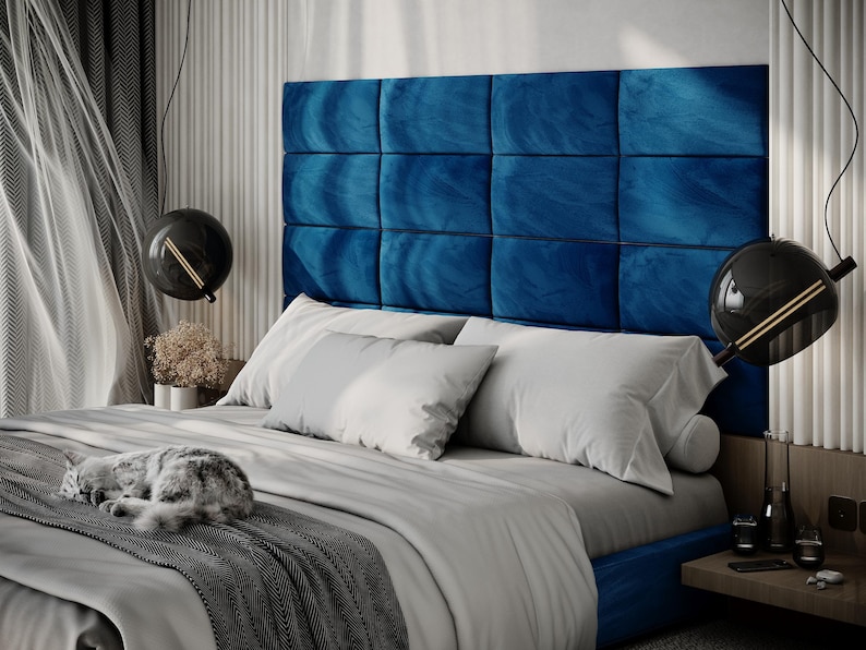 May include: A modern bedroom featuring a bed with a large, square, blue velvet headboard. The bed is made with white pillows and a gray comforter. A gray and white cat is sleeping on the bed. Two black globe pendant lights hang from the ceiling.