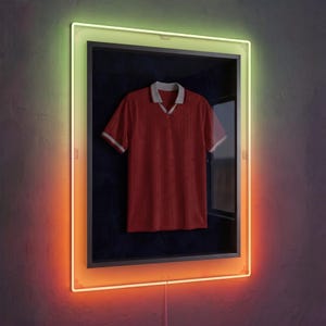 Può includere: Una polo rossa a maniche corte con colletto e bordi delle maniche grigio chiaro, esposta all'interno di una cornice nera rettangolare. La cornice è circondata da un bordo luminoso al neon che sfuma dal verde in alto all'arancione in basso.
