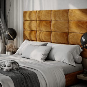 May include: A bedroom scene featuring a bed with a large, tufted, golden-brown headboard. The bed is made with white pillows and a gray comforter. A gray and white cat is curled up on the bed. Two black globe pendant lights hang above the bed.