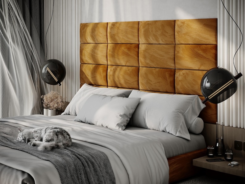 May include: A bedroom scene featuring a bed with a large, tufted, golden-brown headboard. The bed is made with white pillows and a gray comforter. A gray and white cat is curled up on the bed. Two black globe pendant lights hang above the bed.