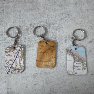 May include: Three rectangular keychains with silver key rings. Each keychain features a different map design. One map is white and blue, another is tan, and the third is white and tan. The keychains are on a gray surface.