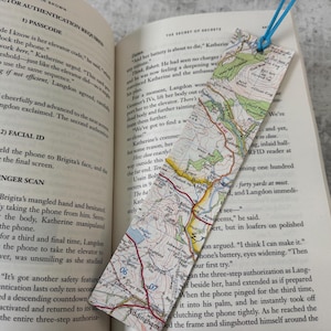 Puede incluir: Un marcapáginas rectangular con un diseño de mapa, con una cuerda azul para una fácil colocación. El marcapáginas está colocado en un libro abierto, mejorando la experiencia de lectura. Los detalles del mapa son visibles.