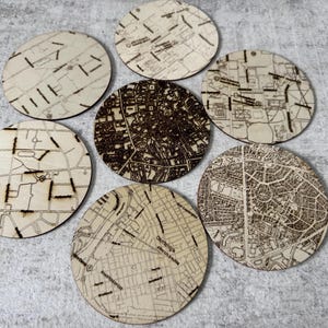 May include: Seven round wooden coasters with detailed map designs. The coasters feature a light wood color with dark brown etched lines depicting city streets and landmarks. A unique set for home decor.