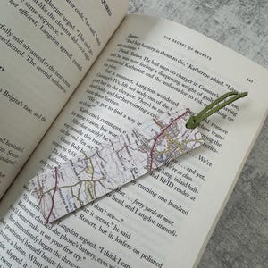 May include: A handmade bookmark featuring a map design with a green suede tassel. The bookmark is placed in an open book with text visible on the pages. The bookmark's map design includes roads and geographical features.