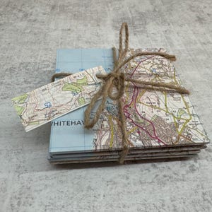 Può includere: Un set di sottobicchieri quadrati con dettagliate mappe, legati con spago. I sottobicchieri hanno uno sfondo azzurro con dettagli colorati della mappa. È attaccata una piccola etichetta con dettagli della mappa.