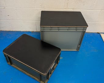 May include: Two gray storage containers with black tops are positioned on a blue floor. One container is upright, while the other lies on its side. The containers are made of a ribbed plastic material.