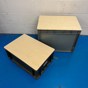 May include: Two grey plastic storage crates with light wood-effect lids. One crate is upright, and the other is on its side. The crates are on a blue floor, with a white brick wall in the background.