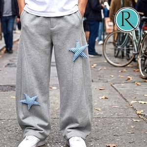 Op de afbeelding: Grijze joggingbroek met twee blauw-wit gestreepte sterapplicaties. De broek met wijde pijpen heeft een naad aan de voorkant en wordt gedragen met witte sneakers. De foto is buiten op straat gemaakt.