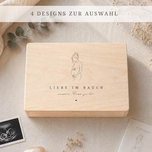 Op de afbeelding: Een lichtgekleurde houten kist met een illustratie van een zwangere vrouw en de tekst "LIEBE IM BAUCH" en "unsere Reise zu dir". De afbeelding bevat ook een echo foto en een kaart met een vergelijkbare illustratie.