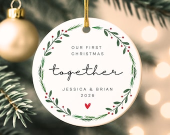May include: A round white ceramic ornament hanging by a gold string. The center features the text "OUR FIRST CHRISTMAS together JESSICA & BRIAN 2026" with a small red heart. A green pine wreath with red berries encircles the printed text.