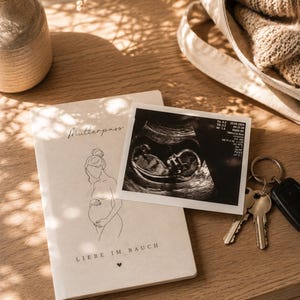 Op de afbeelding: Een stilleven met een notitieboek met het woord "Mutterpass" en een lijntekening van een zwangere persoon. Een echografie en sleutels zijn ook aanwezig, op een houten oppervlak.