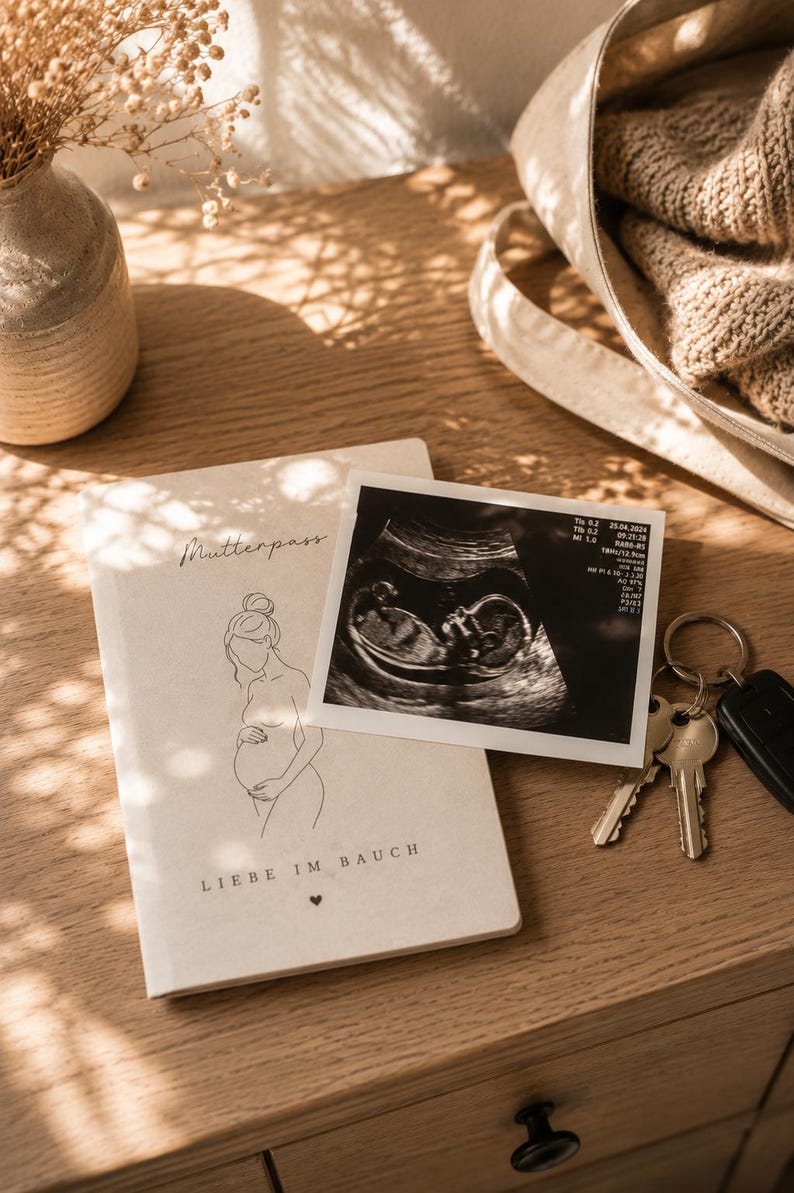 Op de afbeelding: Een stilleven met een notitieboek met het woord "Mutterpass" en een lijntekening van een zwangere persoon. Een echografie en sleutels zijn ook aanwezig, op een houten oppervlak.