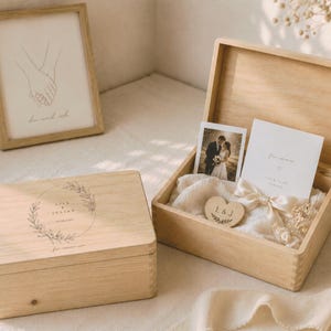 May include: Two wooden boxes, one closed and one open, with wedding-themed contents. The closed box has a circular design with names and a date. The open box contains a photo, card, and heart-shaped wooden piece. A framed drawing is in the background.