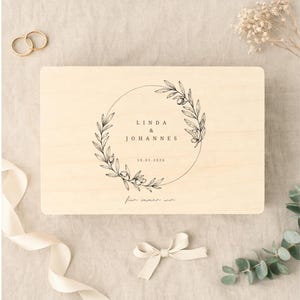 May include: A light-colored wooden box with a circular floral design and the names "LINDA & JOHANNES" and the date "10.01.2026" in black. The box is decorated with a cream-colored ribbon and a small bow.