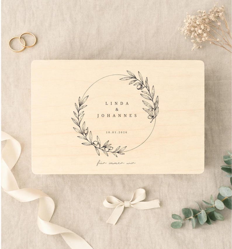 May include: A light-colored wooden box with a circular floral design and the names "LINDA & JOHANNES" and the date "10.01.2026" in black. The box is decorated with a cream-colored ribbon and a small bow.