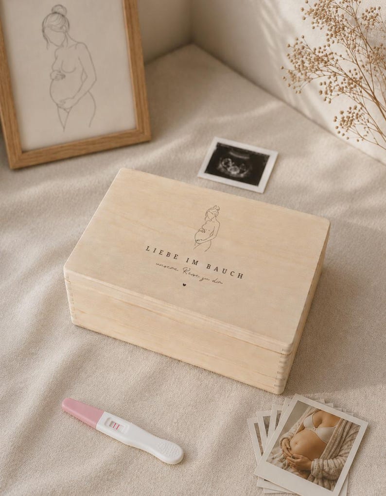 Op de afbeelding: Een lichtgekleurde houten kist met de tekst "LIEBE IM BAUCH" en een lijntekening van een zwangere persoon. De kist staat naast een zwangerschapstest, een ingelijste schets van een zwangere persoon en echografiebeelden.