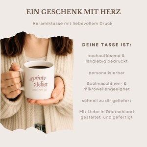 May include: A white ceramic mug filled with coffee, held by a person. The mug features the text "printy atelier" in a pink script font. The image also includes text in German: "EIN GESCHENK MIT HERZ" and "DEINE TASSE IST:"