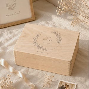 May include: A light-colored wooden box with a lid, featuring a circular floral design with the names "Lisa & Julian" and a date. A framed drawing of hands holding each other and a small photo of a couple are also visible.