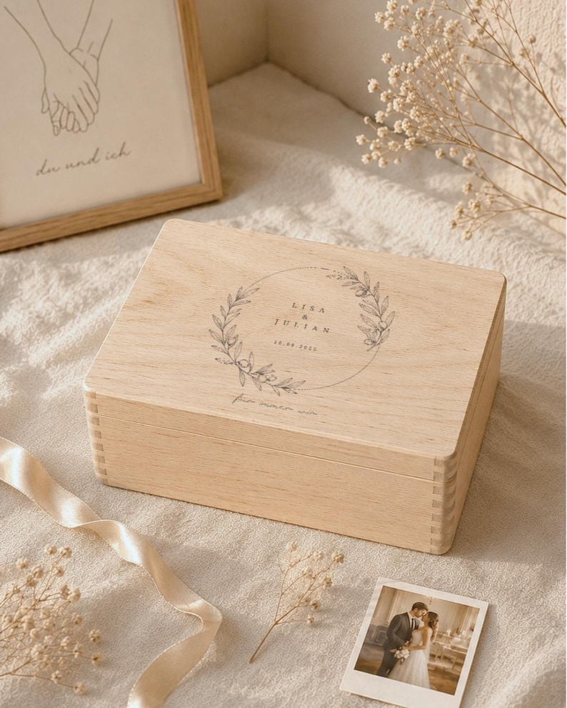 May include: A light-colored wooden box with a lid, featuring a circular floral design with the names "Lisa & Julian" and a date. A framed drawing of hands holding each other and a small photo of a couple are also visible.