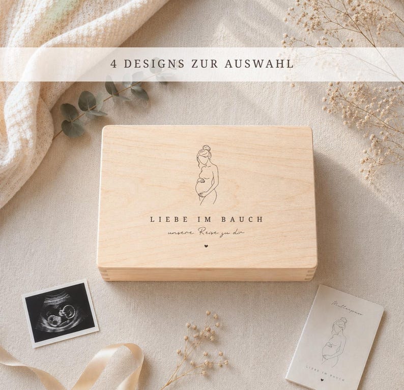 Op de afbeelding: Een lichtgekleurde houten kist met een illustratie van een zwangere vrouw en de tekst "LIEBE IM BAUCH" en "unsere Reise zu dir". De afbeelding bevat ook een echo foto en een kaart met een vergelijkbare illustratie.