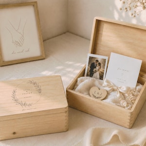 May include: Two wooden boxes, one closed and one open, with wedding-themed items. The open box contains a photo, card, and wooden heart. The closed box has a circular design with text. A framed line drawing of hands holding is in the background.
