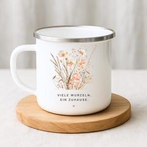 May include: White enamel mug with a silver rim, featuring a floral design in soft pinks and yellows. The mug has a handle and text that reads "VIELE WURZELN. EIN ZUHAUSE." on the front. It sits on a round wooden coaster.