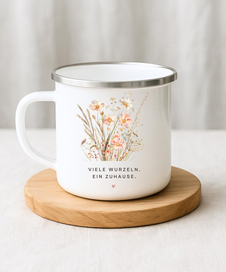 May include: White enamel mug with a silver rim, featuring a floral design in soft pinks and yellows. The mug has a handle and text that reads "VIELE WURZELN. EIN ZUHAUSE." on the front. It sits on a round wooden coaster.
