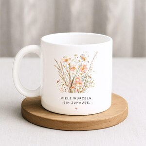 May include: White ceramic mug featuring a floral design with pink and cream-colored flowers, green leaves, and a small ladybug. The mug has the text "VIELE WURZELN. EIN ZUHAUSE." and a small heart. The mug sits on a round wooden coaster.