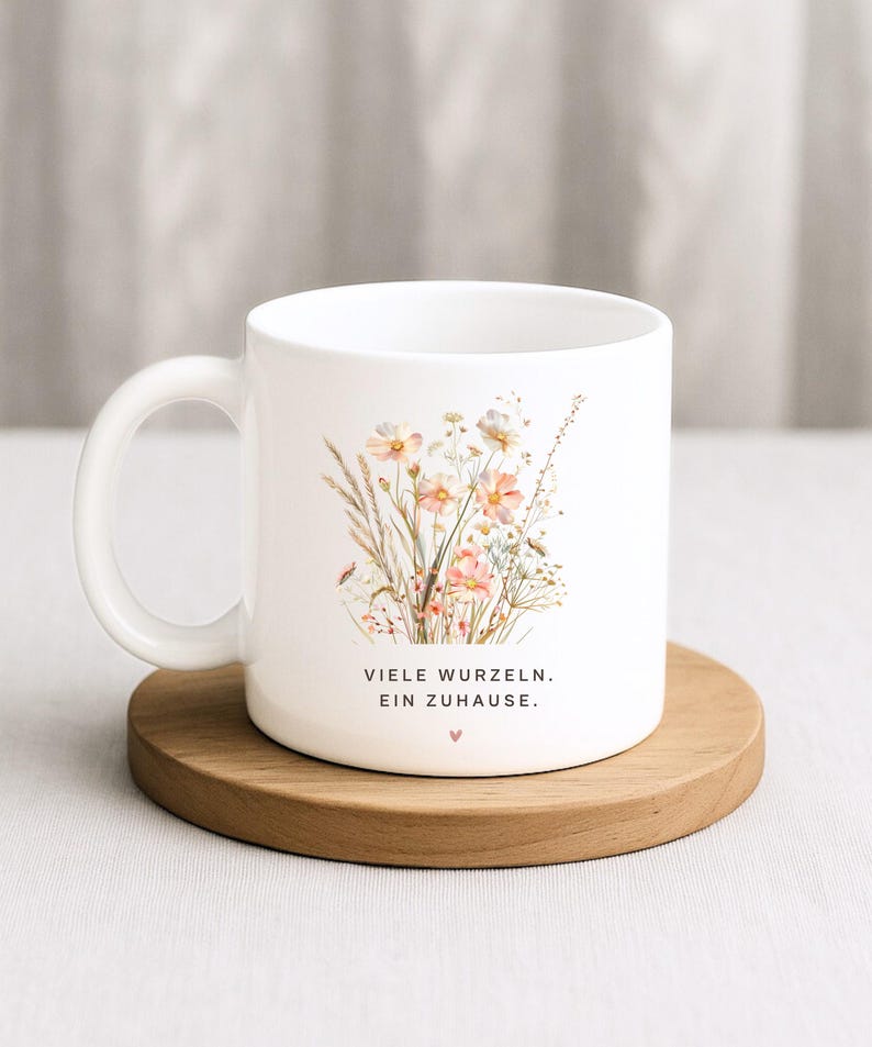 May include: White ceramic mug featuring a floral design with pink and cream-colored flowers, green leaves, and a small ladybug. The mug has the text "VIELE WURZELN. EIN ZUHAUSE." and a small heart. The mug sits on a round wooden coaster.