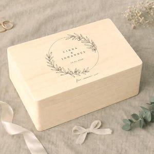 May include: A light-colored wooden box with a hinged lid, featuring a circular floral design and text. The box is rectangular and appears to be for keepsakes or a gift. Gold rings and a white ribbon are also visible.