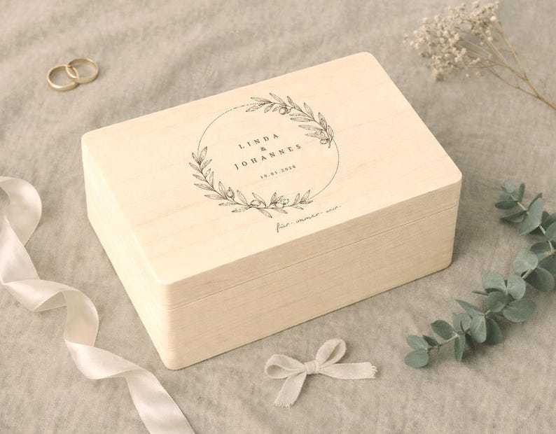 May include: A light-colored wooden box with a hinged lid, featuring a circular floral design and text. The box is rectangular and appears to be for keepsakes or a gift. Gold rings and a white ribbon are also visible.