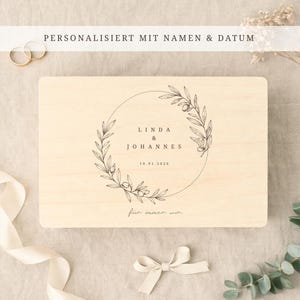 May include: A light-colored wooden box with a floral wreath design and the names "Linda & Johannes" and the date "10.01.2026" printed on it. The text "PERSONALISIERT MIT NAMEN & DATUM" is at the top.