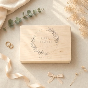 May include: A light-colored wooden box with a circular floral design and the names "LINDA & JOHANNES" and the date "10.01.2016" engraved on the top. The box is surrounded by wedding-themed decorations, including gold rings and a ribbon.