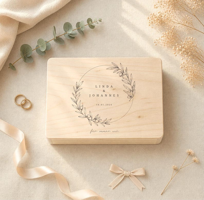 May include: A light-colored wooden box with a circular floral design and the names "LINDA & JOHANNES" and the date "10.01.2016" engraved on the top. The box is surrounded by wedding-themed decorations, including gold rings and a ribbon.